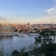 Ein Blick auf Prag mit traditionellen Dächern und modernen Gebäuden, flankiert von einem breiten Fluss und grünen Bäumen im Vordergrund.