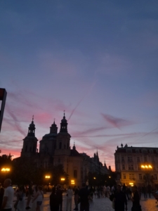 Silhouette der Teyn-Kirche und des Altstädter Rathauses vor einem farbenfrohen Abendhimmel. Menschen gehen auf dem belebten Platz bei eingeschaltem Straßenlaternenlicht.